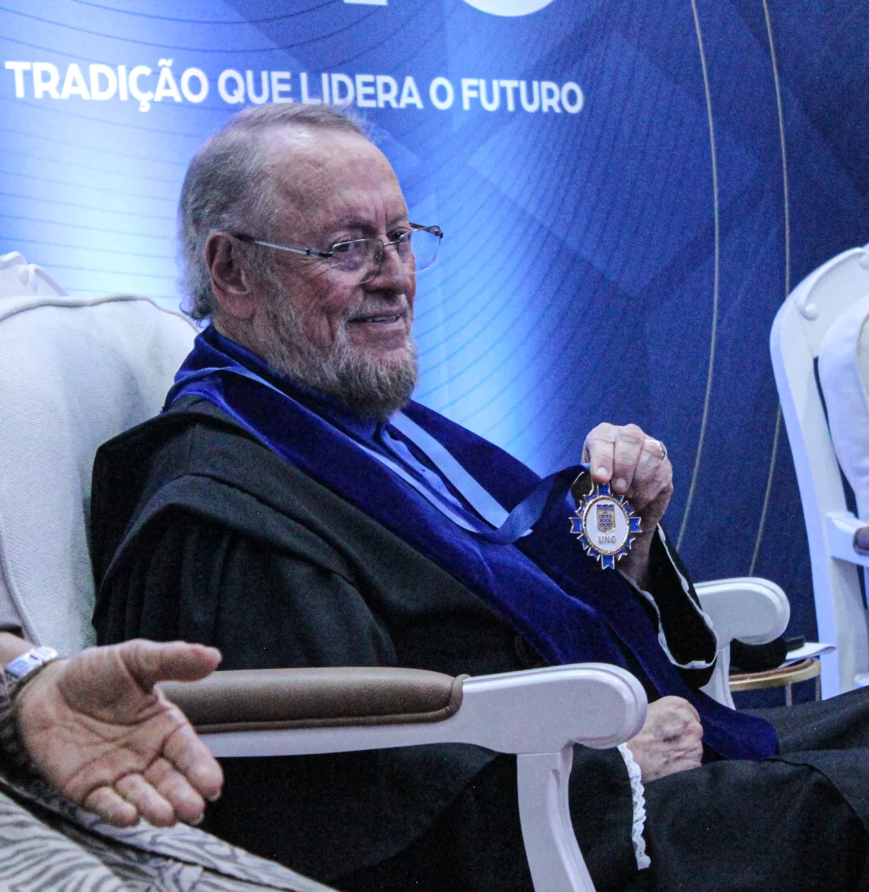 Homem com vestes acadêmicas segurando uma medalha da UNG, diante de um painel azul.